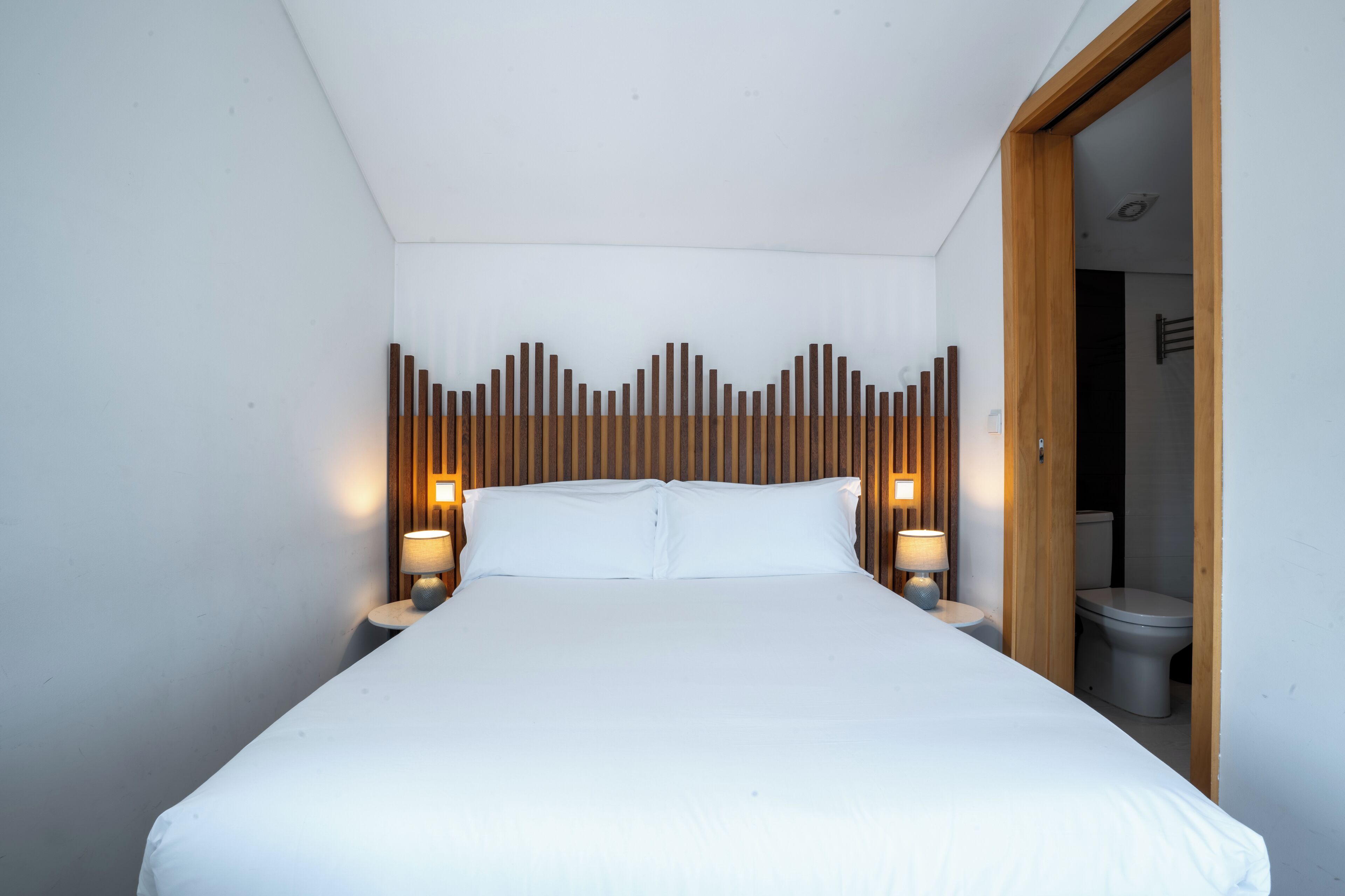 Una cama bien hecha con sábanas blancas, dos lámparas de noche y un cabecero de madera con un diseño original.