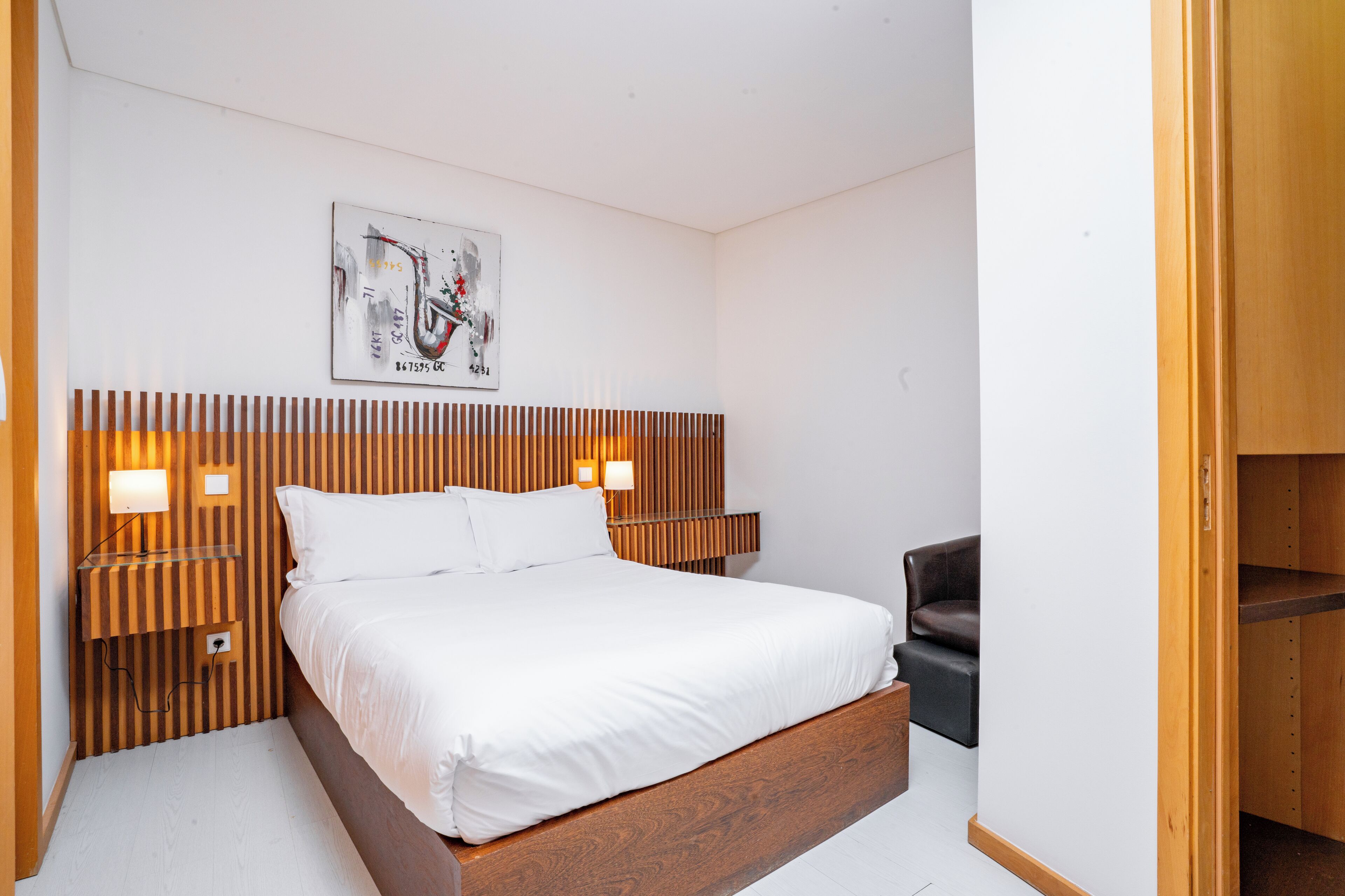 A neatly arranged bedroom with a wooden headboard, a bed with white linens, a black chair, and a wall-mounted artwork.