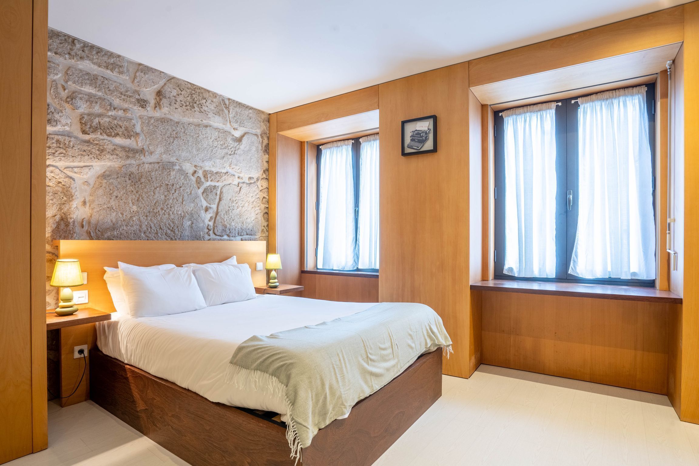 A bedroom with a stone wall, wooden paneling, a large bed, two bedside tables with lamps, and windows with curtains.