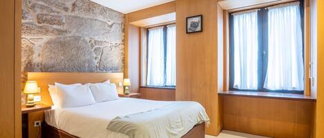 A bedroom with a stone wall, wooden paneling, a large bed, two bedside tables with lamps, and windows with curtains.