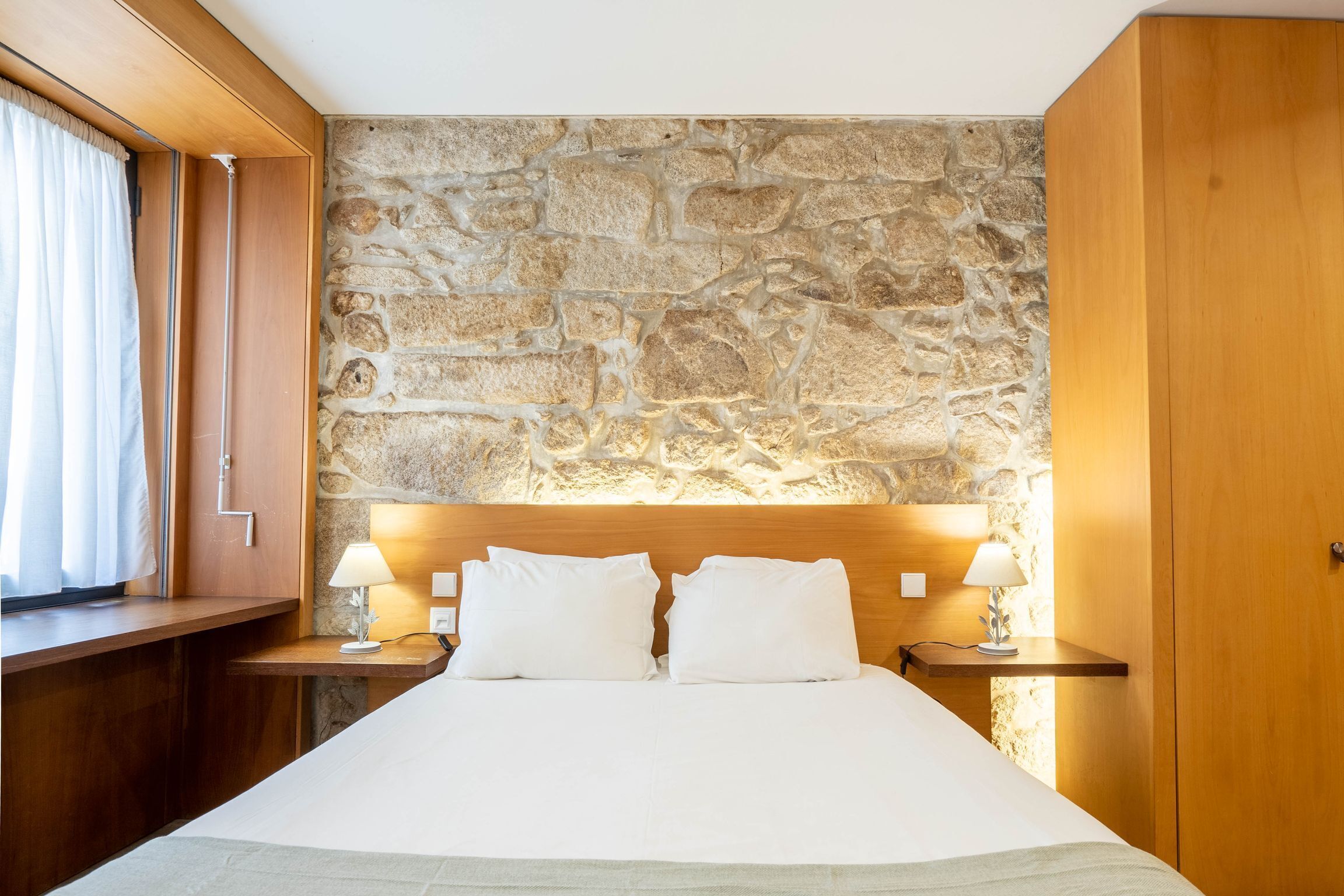 A hotel room with a stone wall, wooden headboard, two bedside tables with lamps, and a large bed with white linens.