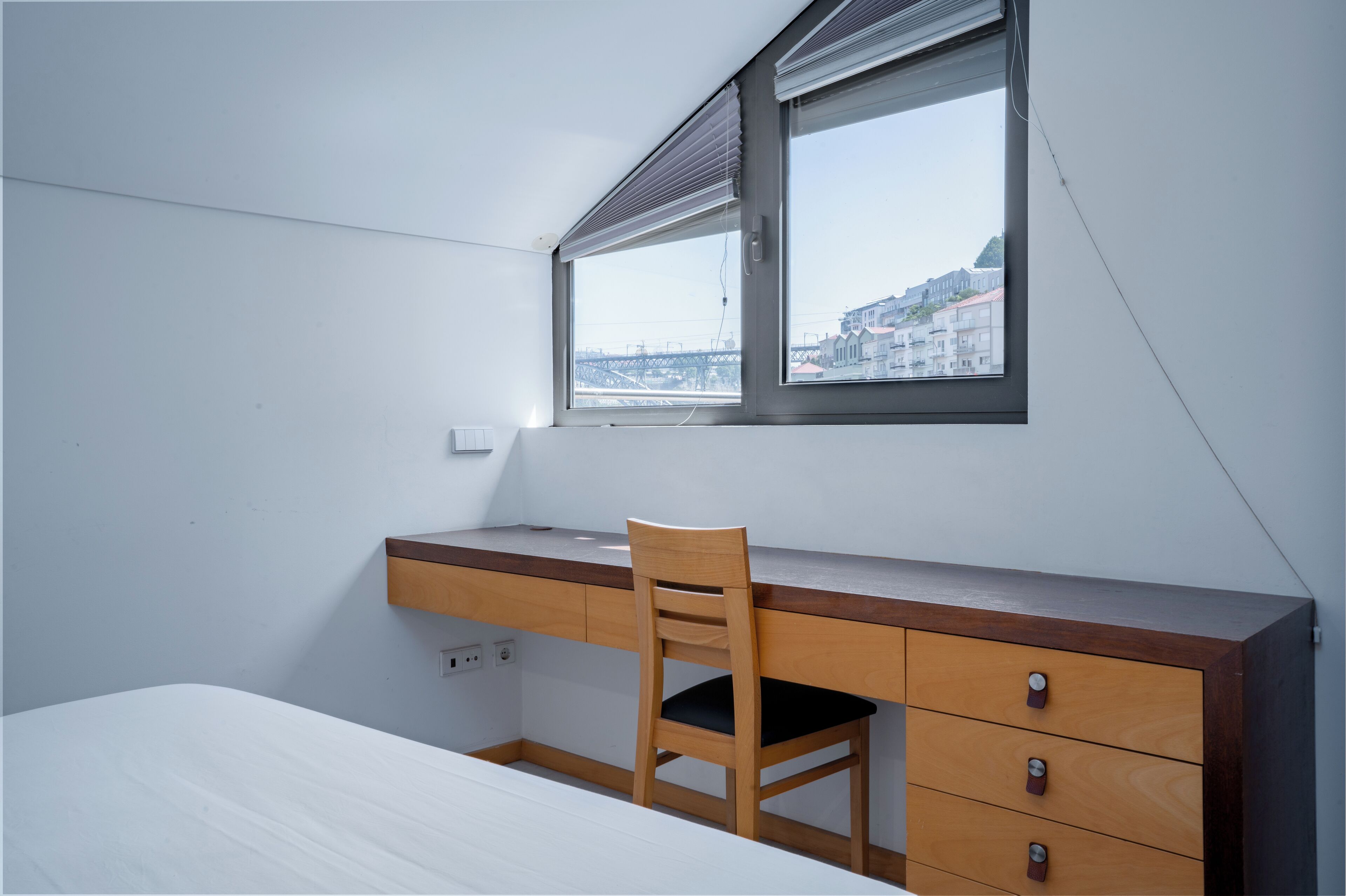 Un dormitorio con cama, escritorio, silla y ventana con vistas a edificios.