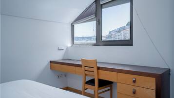 A bedroom with a bed, a desk, a chair, and a window with a view of buildings.