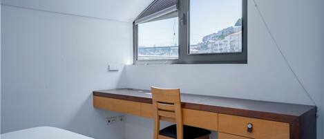 Un dormitorio con cama, escritorio, silla y ventana con vistas a edificios.
