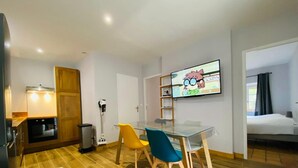 Una habitación de hotel compacta con kitchenette, un comedor con mesa y sillas de vidrio, un televisor montado en la pared y una cama con ventana.