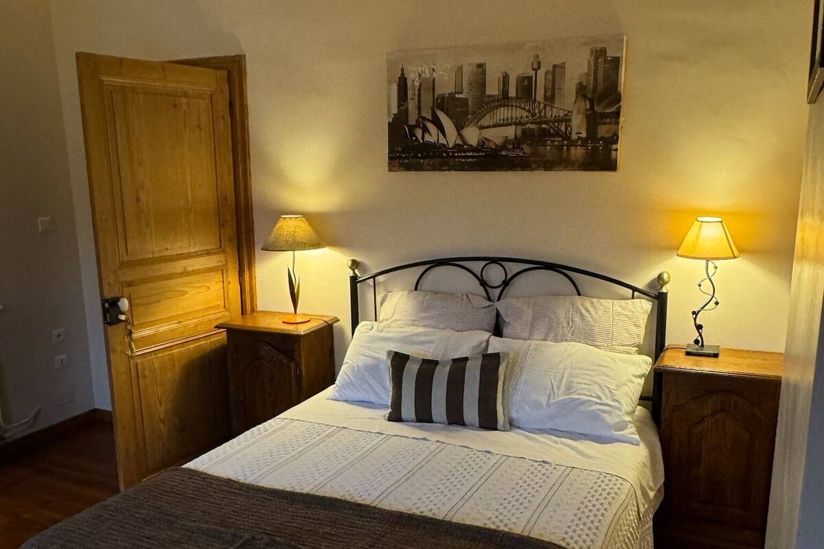 Un dormitorio acogedor con una cama, dos mesitas de noche, una puerta de madera y un cuadro de un paisaje urbano en la pared.