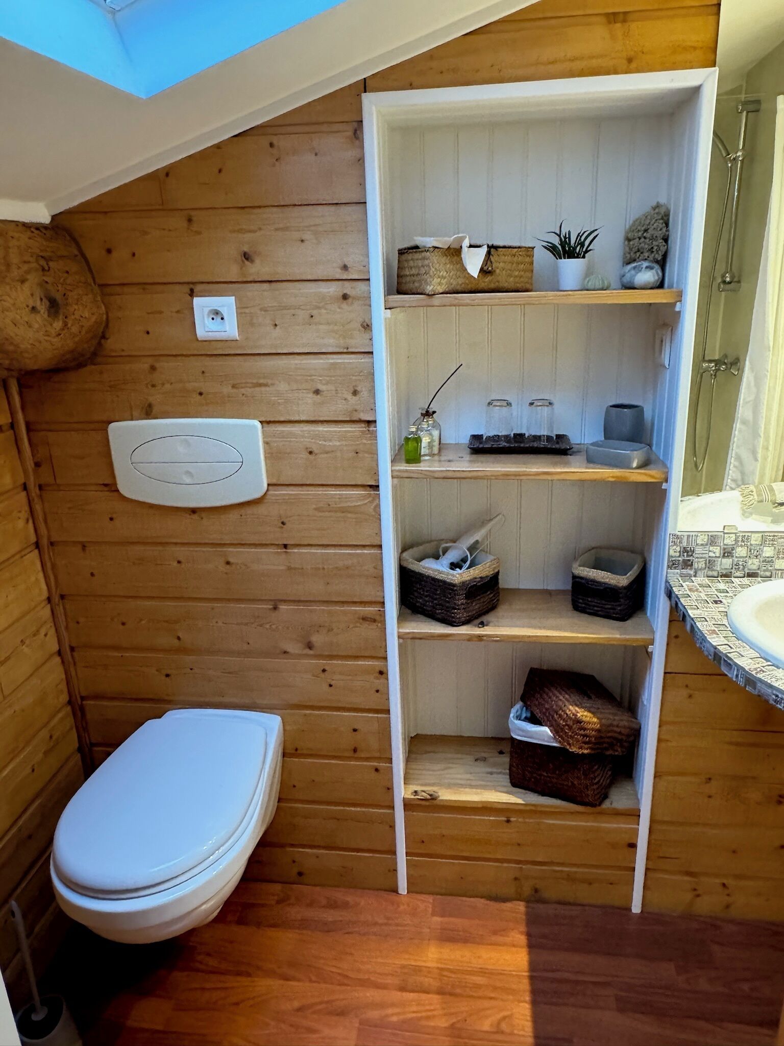 Un baño de madera con tragaluz, inodoro, estantes con cestas y zona de ducha.