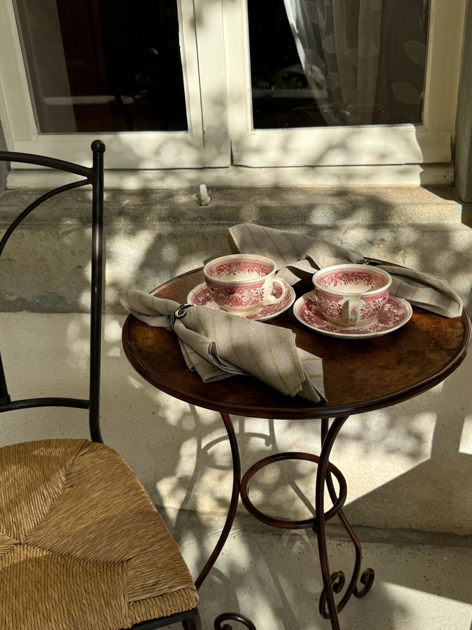 A round wooden table with two matching red and white patterned cups and saucers, a beige cloth napkin, and a metal chair with a woven seat.