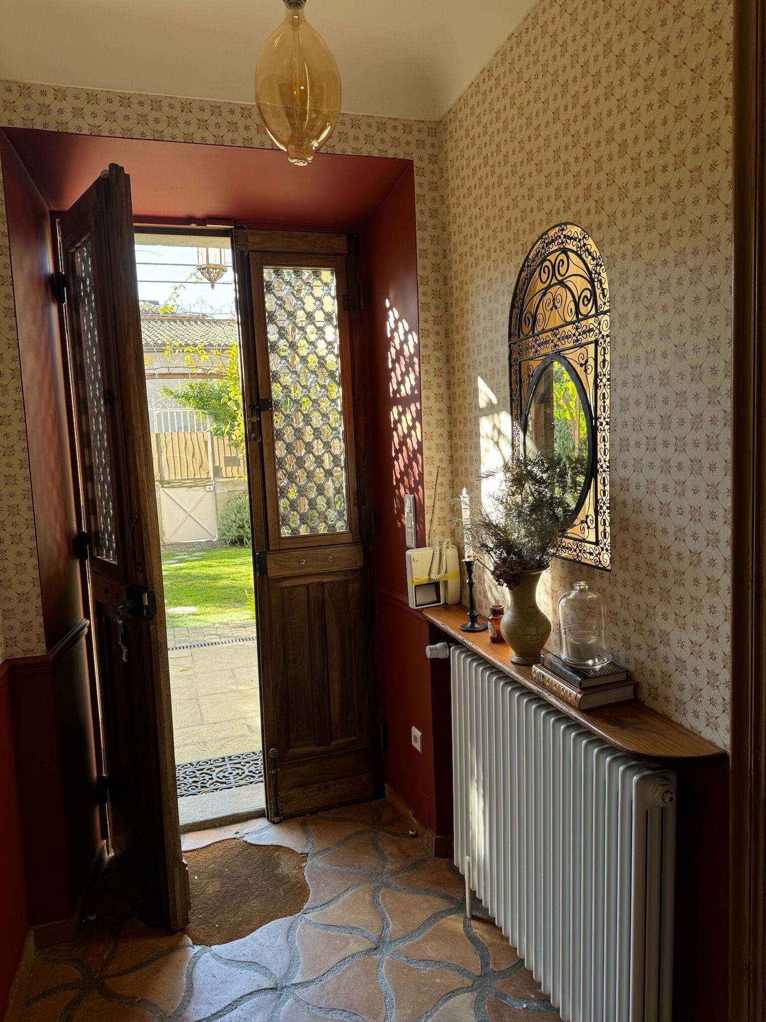 Una habitación con puerta de madera que da a un jardín, un radiador, un espejo y una pequeña mesa con un jarrón y libros.