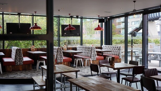 Un espace de restauration intérieur avec des tables et des bancs en bois, de grandes fenêtres donnant sur un paysage verdoyant et des bâtiments, ainsi que des lampes suspendues rouges.