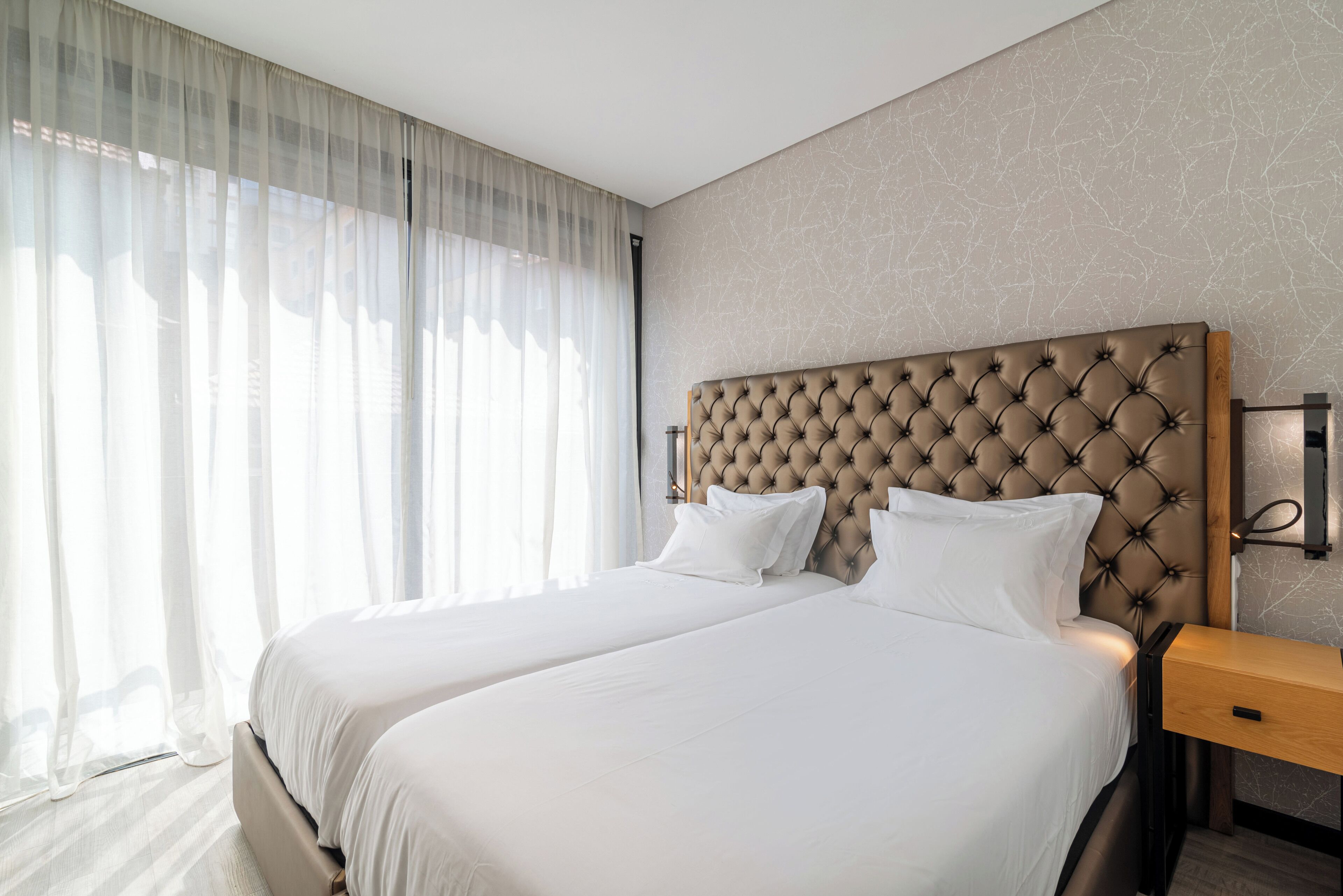 A hotel room with two beds, a wooden nightstand, and a large window with sheer curtains.