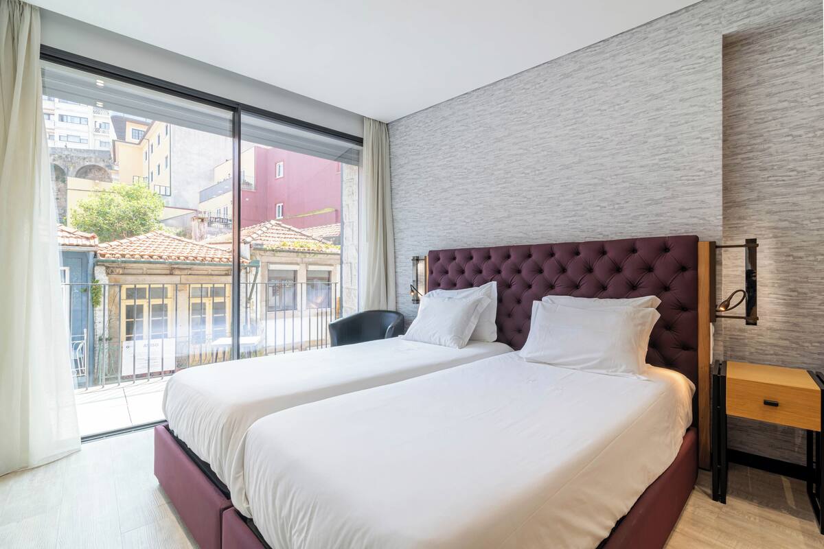Quarto de hotel com uma cama grande, uma mesinha de cabeceira e vista da cidade por uma janela ampla.