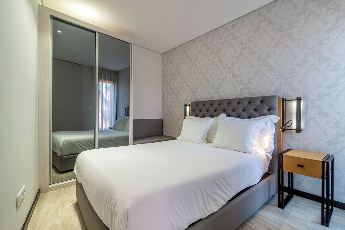 Quarto de hotel moderno com uma cama grande, uma mesa de cabeceira e um guarda-roupa com portas deslizantes.