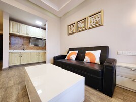 Uma sala de estar moderna com um sofá de couro preto, uma mesa de centro branca e uma área de cozinha com armários de cor clara e painel de azulejos.