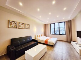 Quarto de hotel moderno com sofá de couro preto, mesa de cabeceira branca, cama com um corredor laranja, uma janela grande com vista para a cidade e três obras de arte florais emolduradas na parede.