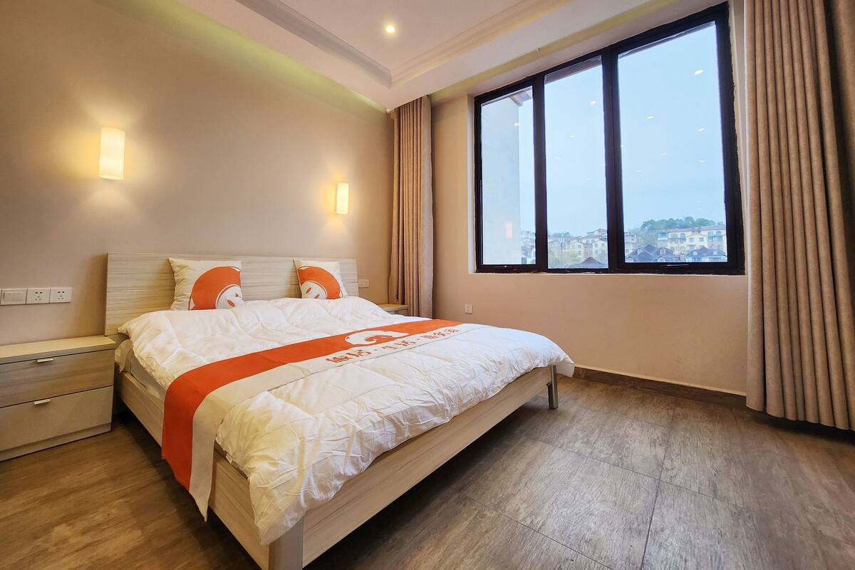 A hotel room with a large bed, a nightstand, a dresser, and a window with a city view.