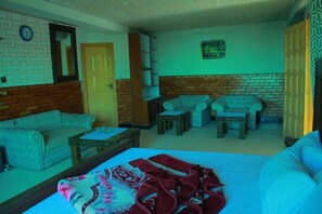 A room with a bed, a sofa, a coffee table, and a small table with a chair. There is a door and a wall with a clock and a framed picture.