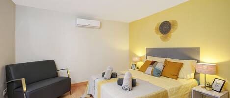 A hotel room with a bed, a sofa, a bedside table, and an air conditioning unit.