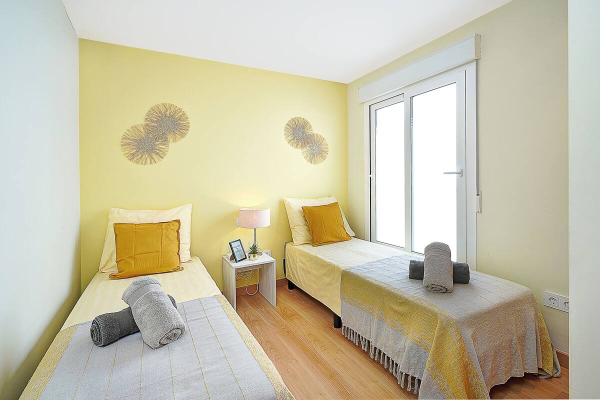 Une chambre avec deux lits, chacun agrémenté d’un oreiller jaune et d’une serviette roulée. On y trouve une table de chevet avec une lampe et une petite plante. La chambre est dotée d’une fenêtre à cadre blanc.