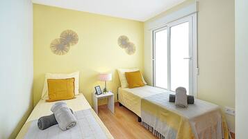 Habitación con dos camas, cada una con una almohada amarilla y una toalla enrollada. Hay una mesita de noche con una lámpara y una planta pequeña. La habitación tiene una ventana con marco blanco.