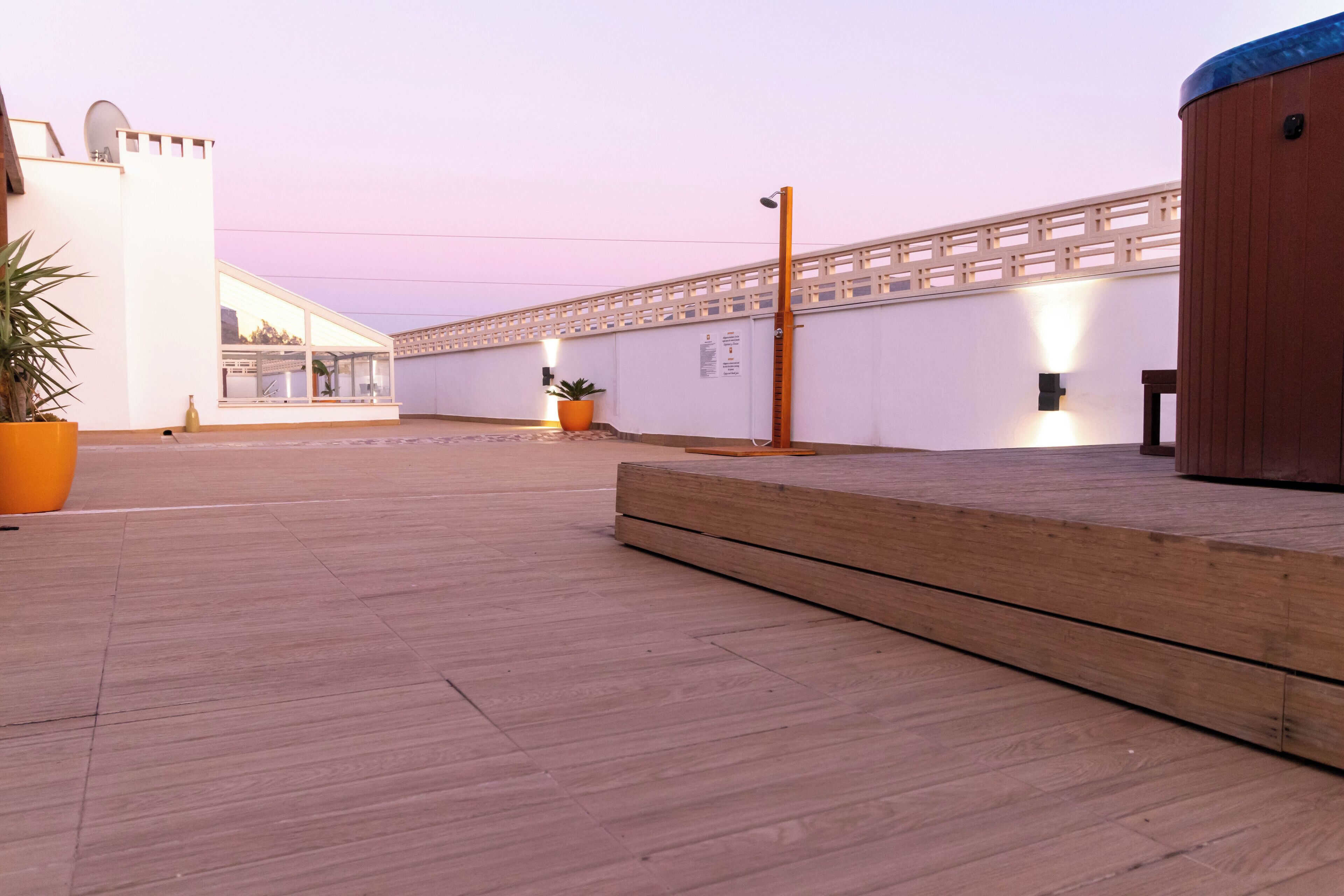 Een ruim buitengebied met een houten terras, potplanten en een wit gebouw met een houten omheining.