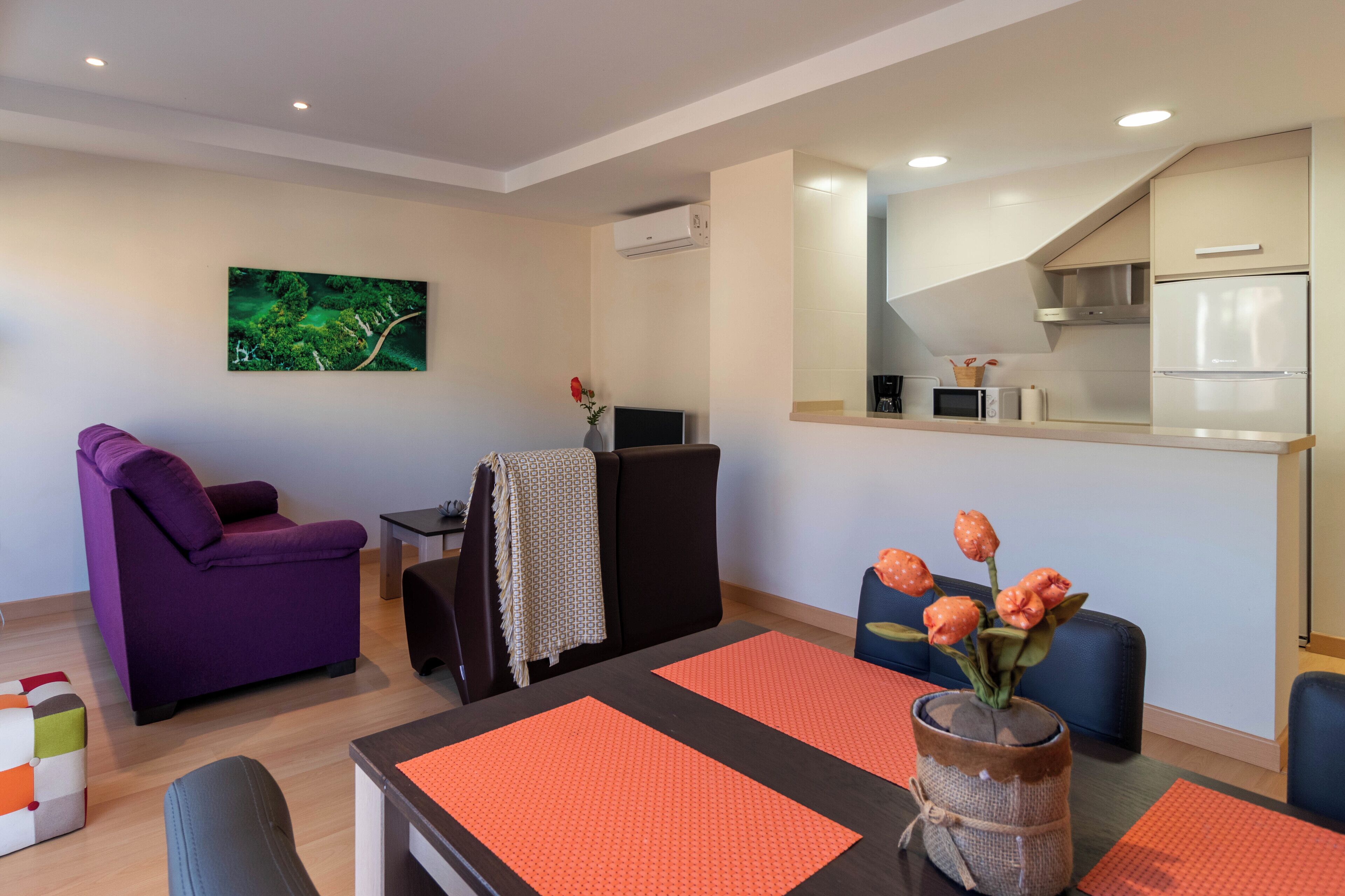 Una sala moderna con un sillón púrpura, una mesa de comedor con individuales anaranjados y una zona de cocina con refrigerador y microondas.