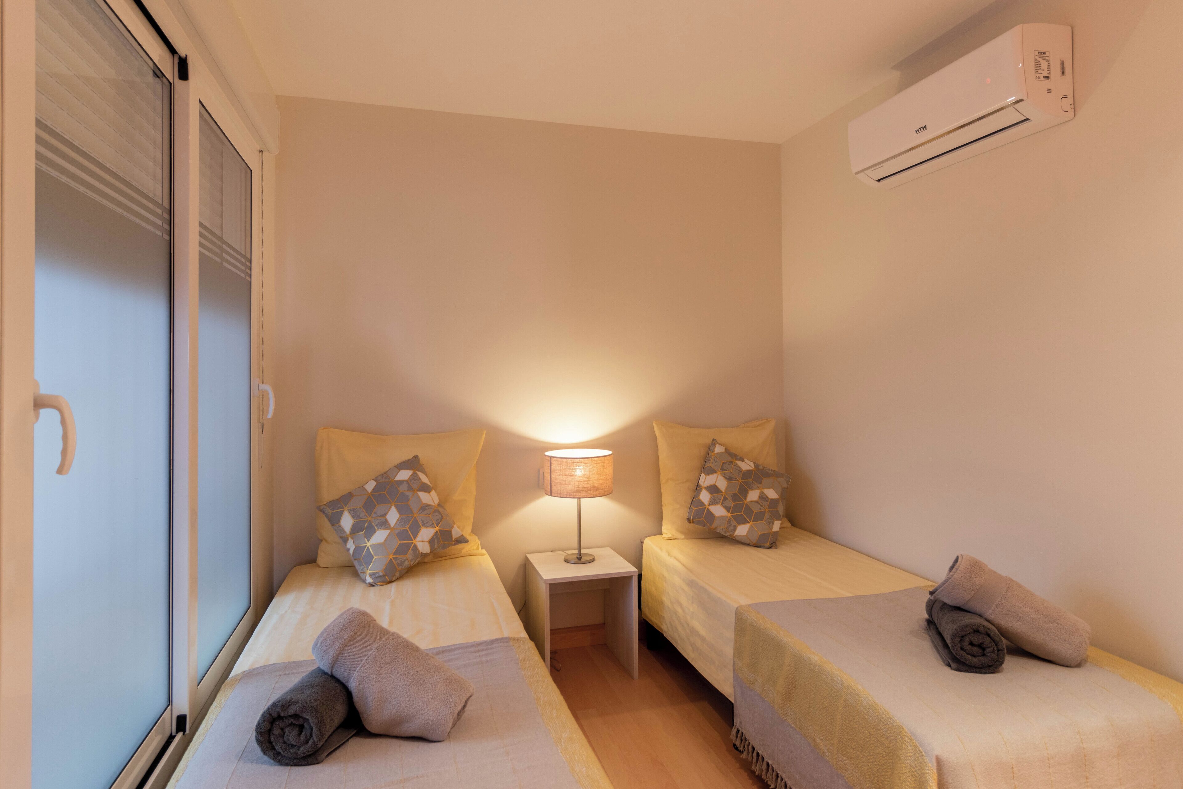 A small, cozy room with two single beds, a nightstand with a lamp, and an air conditioning unit on the wall.