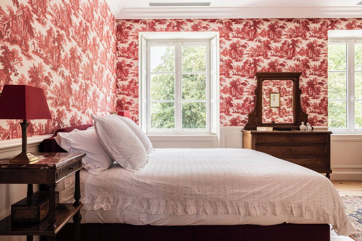 A bedroom with a large bed, a nightstand, a dresser, and a window with a view of trees.
