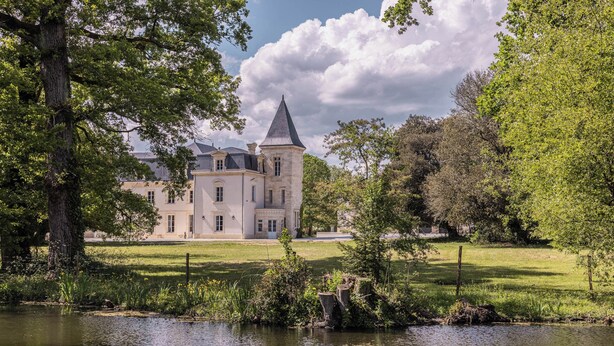 A large, historic building with a prominent tower, surrounded by lush greenery and a calm body of water. - Château Sénéjac (Le Pian-Medoc)