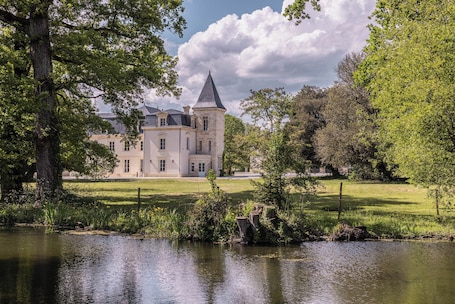 A large, historic building with a prominent tower, surrounded by lush greenery and a calm body of water.