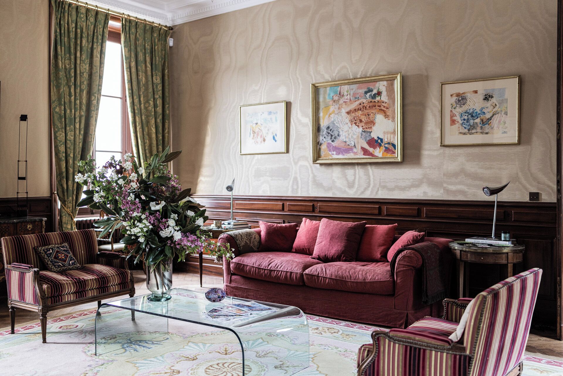 A room with a red sofa, striped armchairs, a glass coffee table, and a large floral arrangement. There are three framed artworks on the wall.