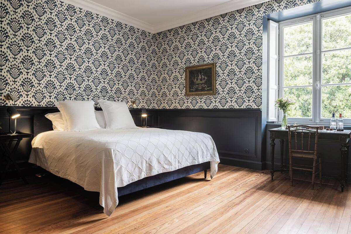 A bedroom with a large bed, dark wooden floor, a window with a view of greenery, and a desk with a vase of flowers.