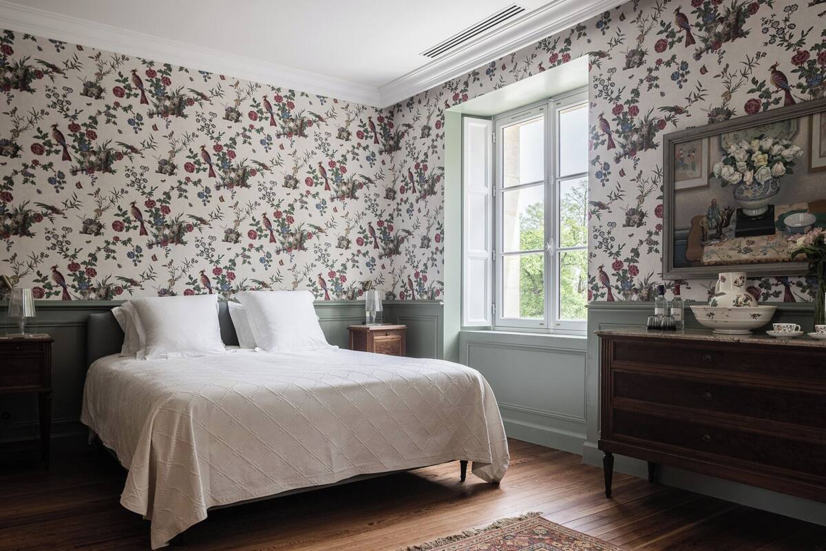 A bedroom with floral wallpaper, a large window, a bed, a dresser, and a painting on the wall.