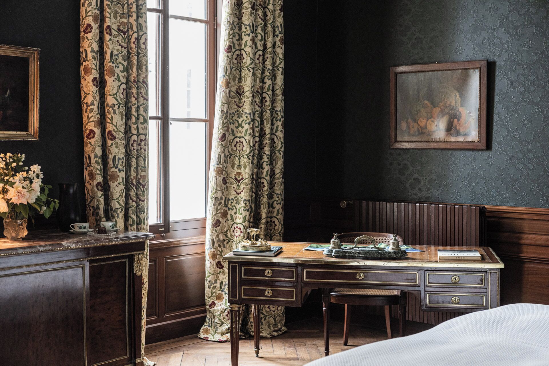 Una habitación con un escritorio de madera, una silla, una cama, un ventanal con cortinas, un cuadro en la pared y un florero con flores sobre el escritorio.