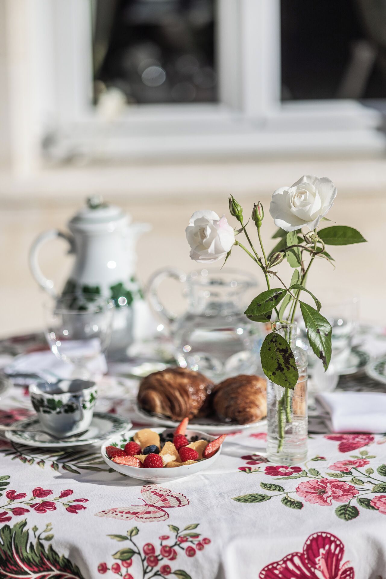 Une table dressée avec une nappe à motifs floraux, une théière, des tasses, une assiette de fruits et un vase contenant des roses blanches.