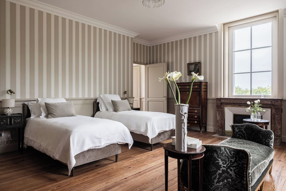 A hotel room with two beds, a fireplace, a large window, and a vase with flowers.