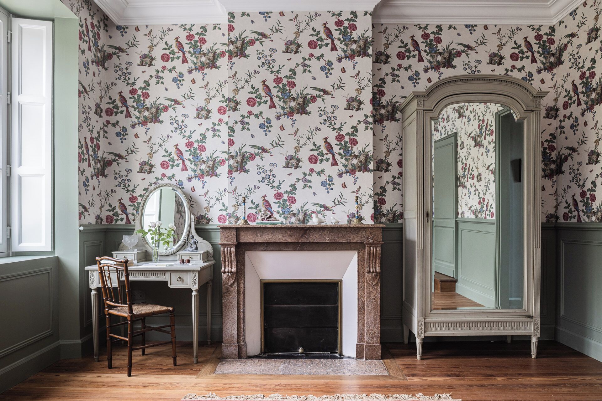 Une pièce avec un papier peint à motifs floraux, une cheminée, un miroir et une chaise en bois.