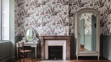 Une pièce avec un papier peint à motifs floraux, une cheminée, un miroir et une chaise en bois.