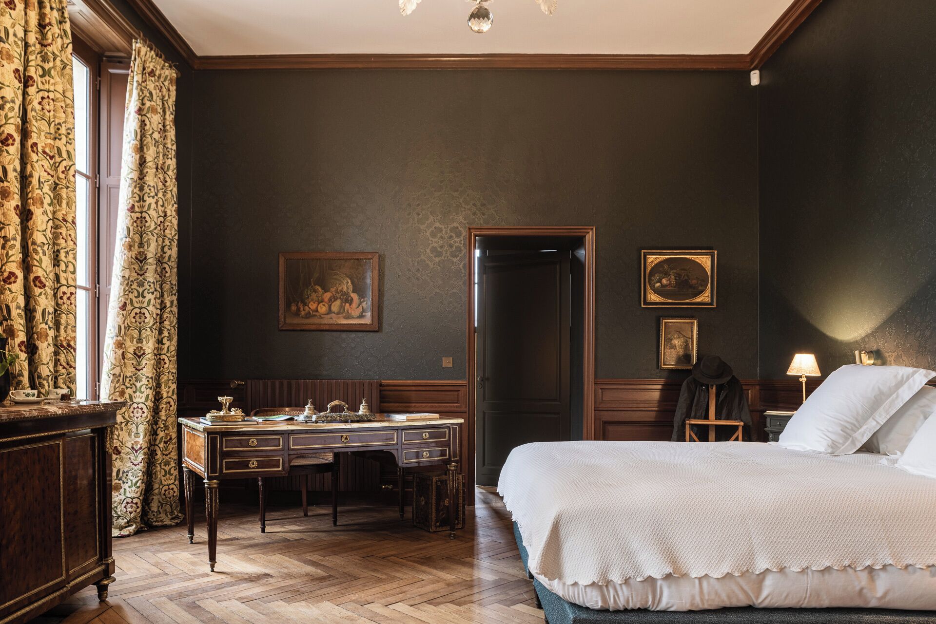 A bedroom with a large bed, a wooden desk, and a chair. There are two framed pictures on the wall, a door, and a window with curtains.