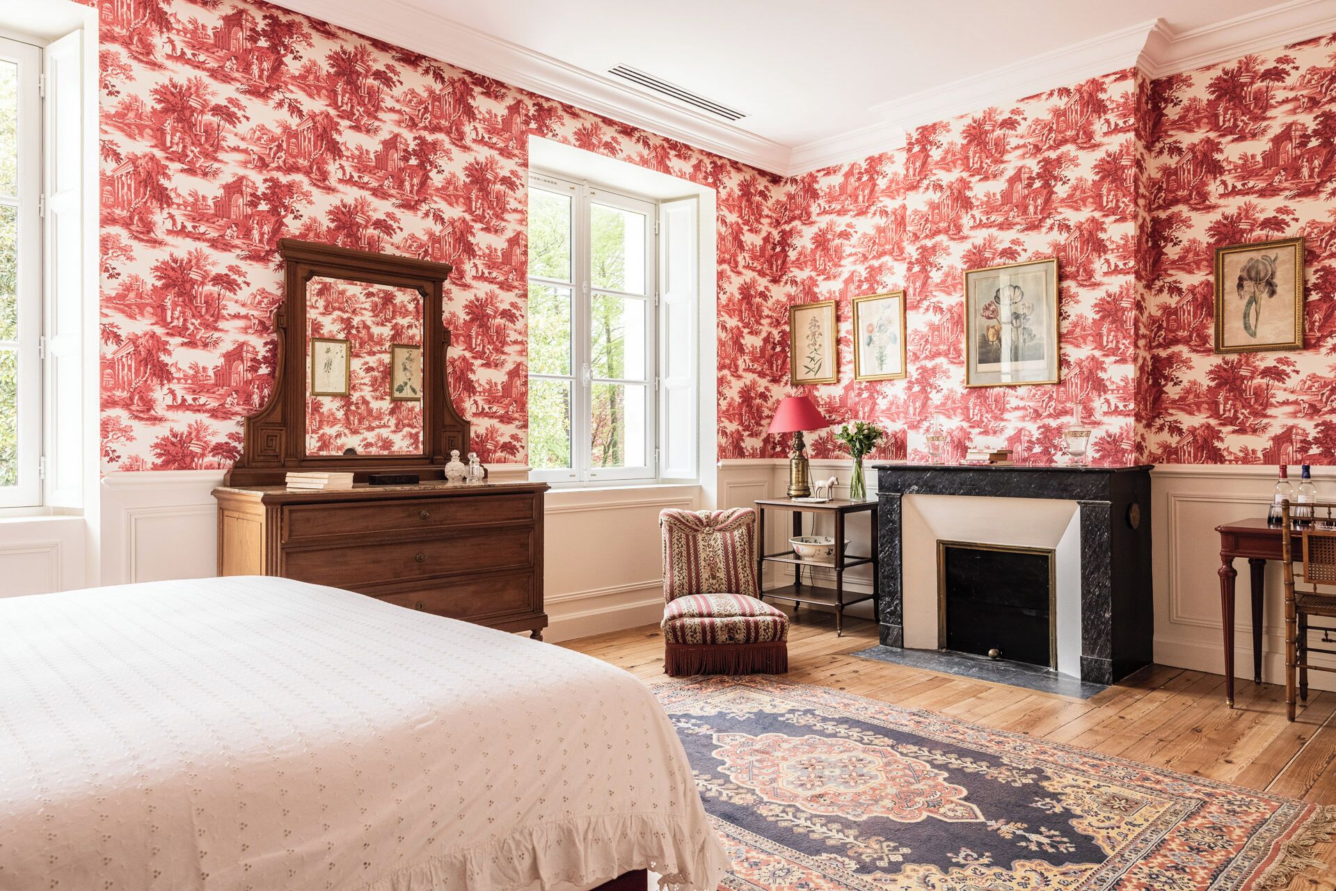 Un dormitorio con papel tapiz floral, chimenea, cama y un ventanal grande.