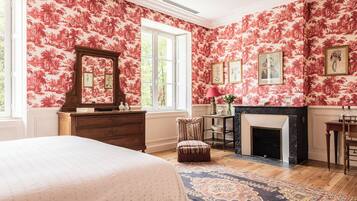 Un dormitorio con papel tapiz floral, chimenea, cama y un ventanal grande.