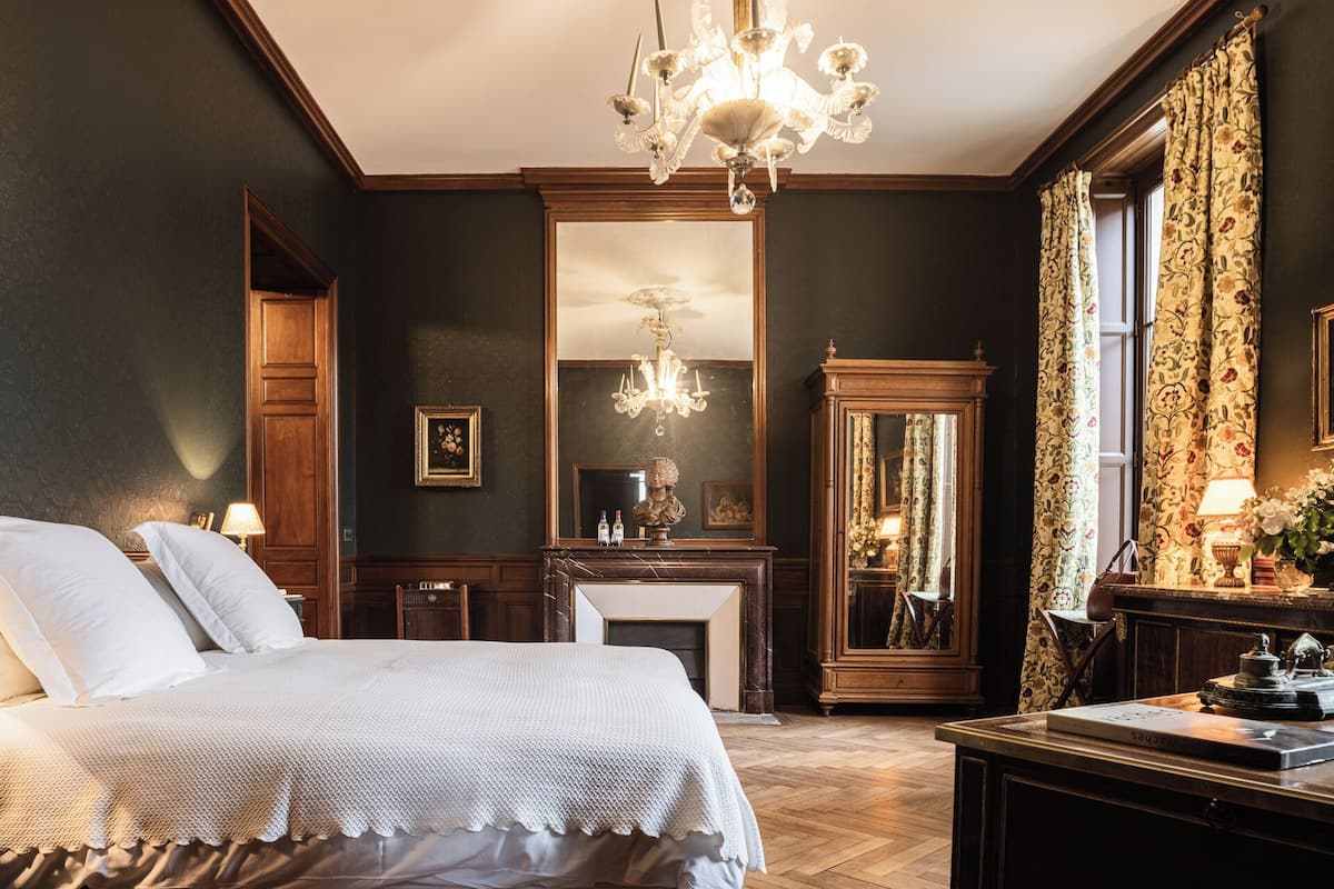 A classic room with a large bed, a fireplace, a mirror, and a chandelier.