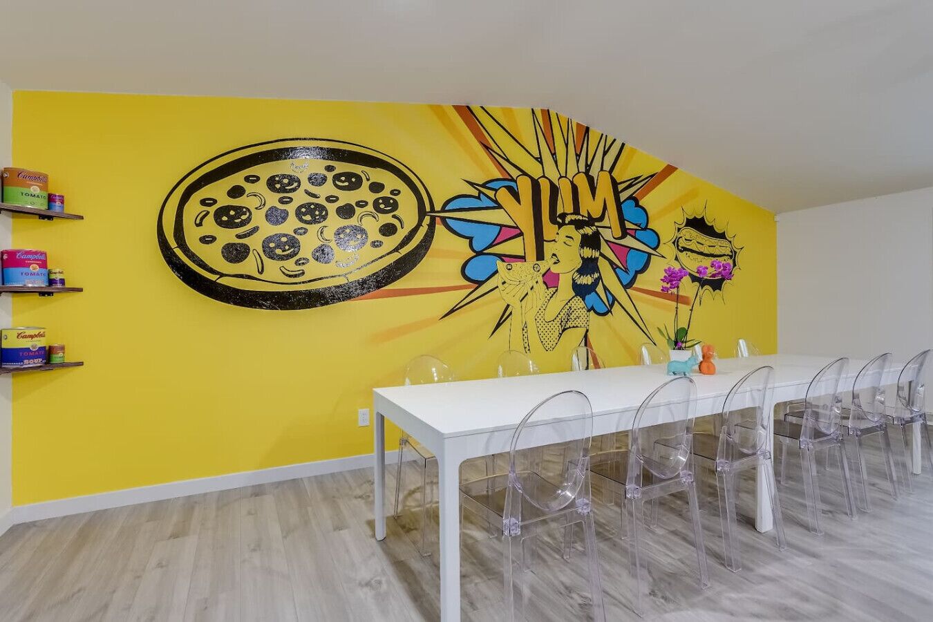 Un espace repas avec une table blanche et des chaises transparentes, un mur jaune orné d’une image de pizza et d’œuvres d’art abstraites, et des étagères remplies de produits alimentaires.