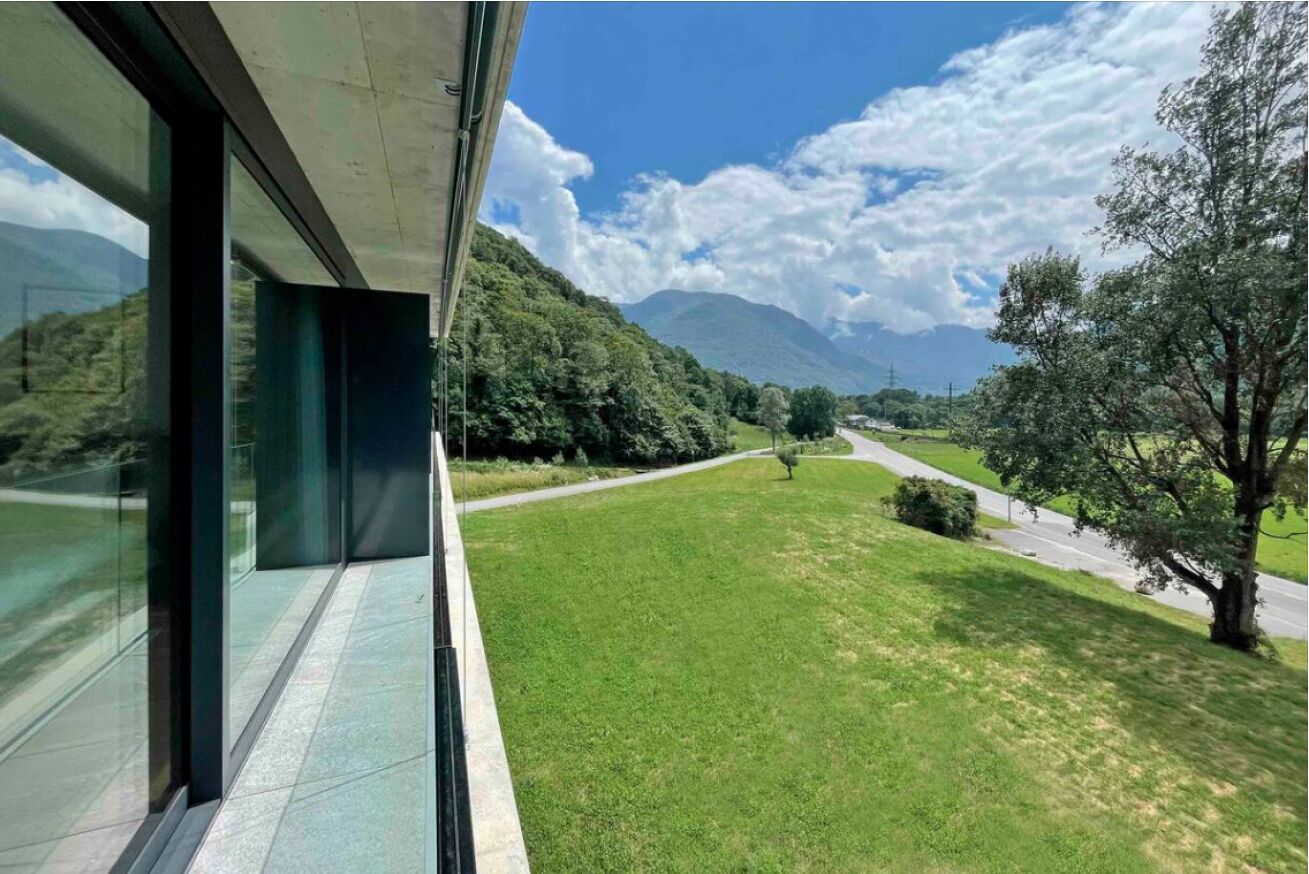 A modern building with large windows overlooking a green landscape with a road and trees.