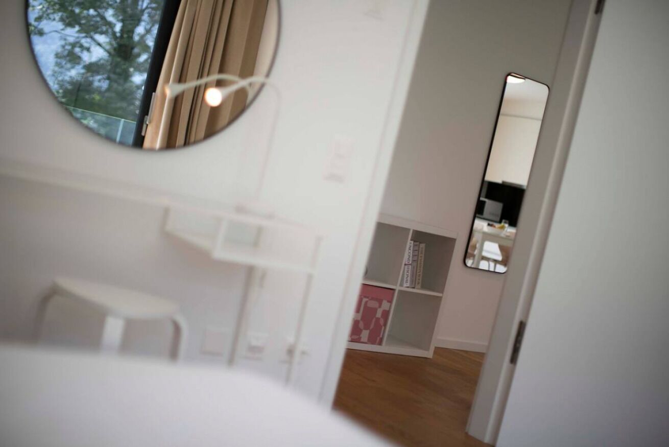 Un dormitorio con un espejo redondo, un taburete blanco pequeño y un armario blanco.
