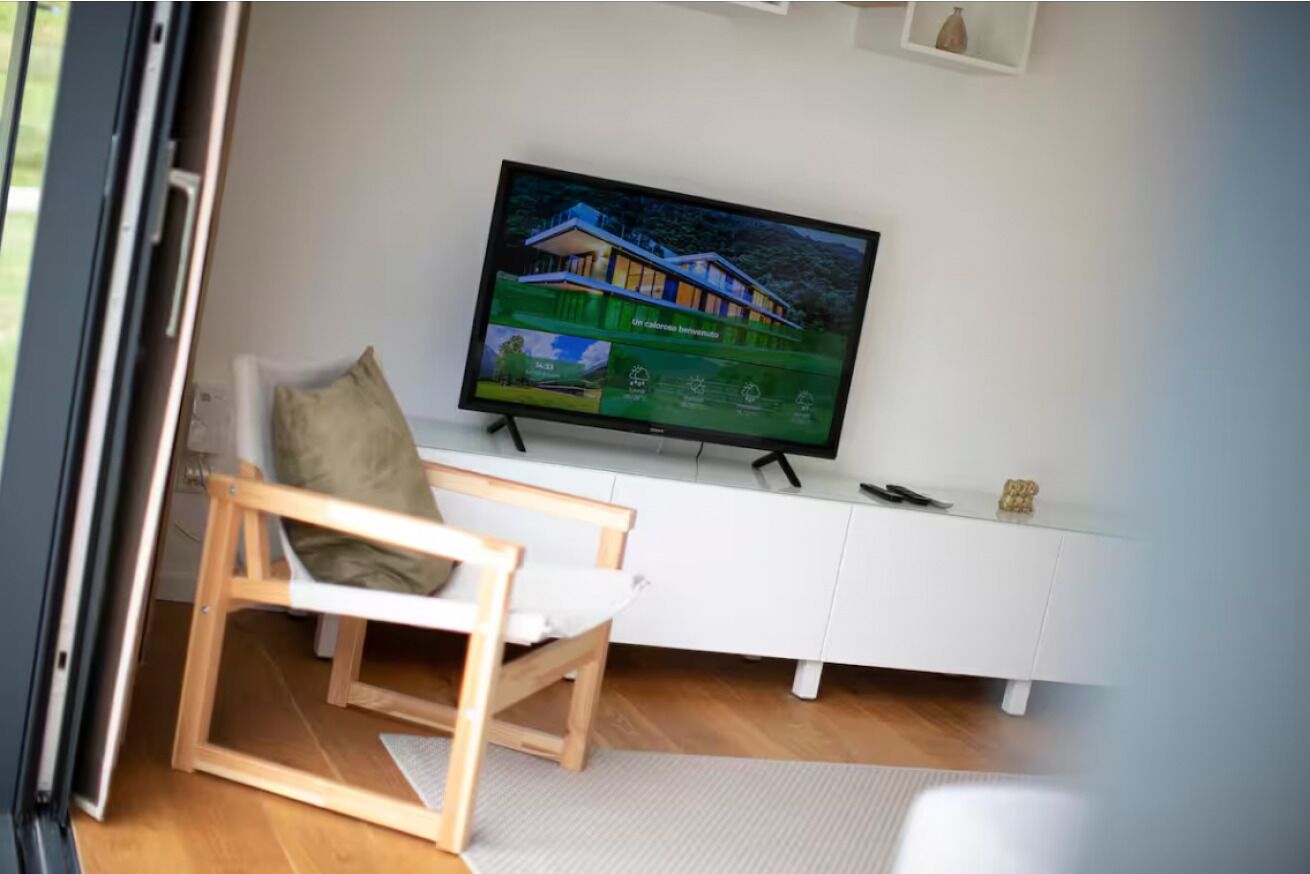 Una habitación con una televisión mostrando una casa, un mueble blanco y una silla de madera.