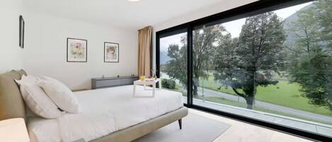 Une chambre moderne avec un grand lit, une table de chevet, une commode et une fenêtre donnant sur des arbres et une route.
