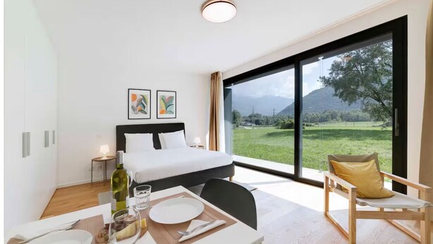 A modern hotel room with a large bed, a dining area with a table set for two, and a view of a green landscape through large windows. - An oasis of peace in nature (Auriga 04 Residenza Ticino) (Bellinzona)