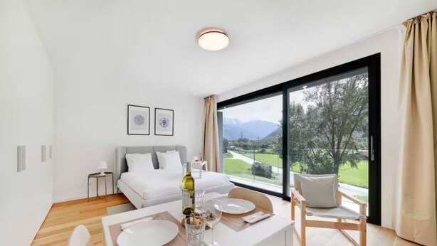 A modern hotel room with a dining area, a bed with white linens, a large window with a view of greenery and mountains, and a small side table with a lamp. - An oasis of peace in nature (Cassiopea 13 Residenza Ticino) (Bellinzona)