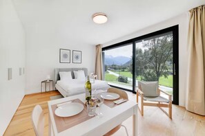 A modern hotel room with a dining area, a bed with white linens, a large window with a view of greenery and mountains, and a small side table with a lamp. - An oasis of peace in nature (Cassiopea 13 Residenza Ticino) (Bellinzona)